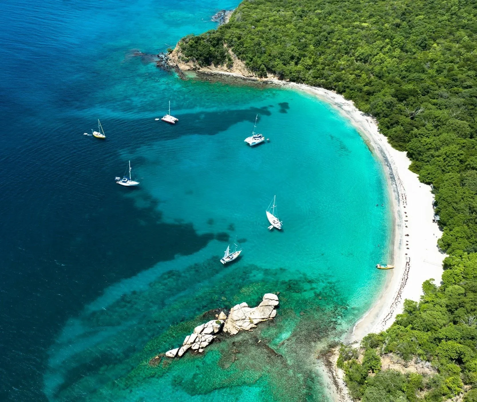 Aerial view of sailing yachts anchored in a turquoise bay beside a white sandy beach and lush tropical coastline in the Caribbean