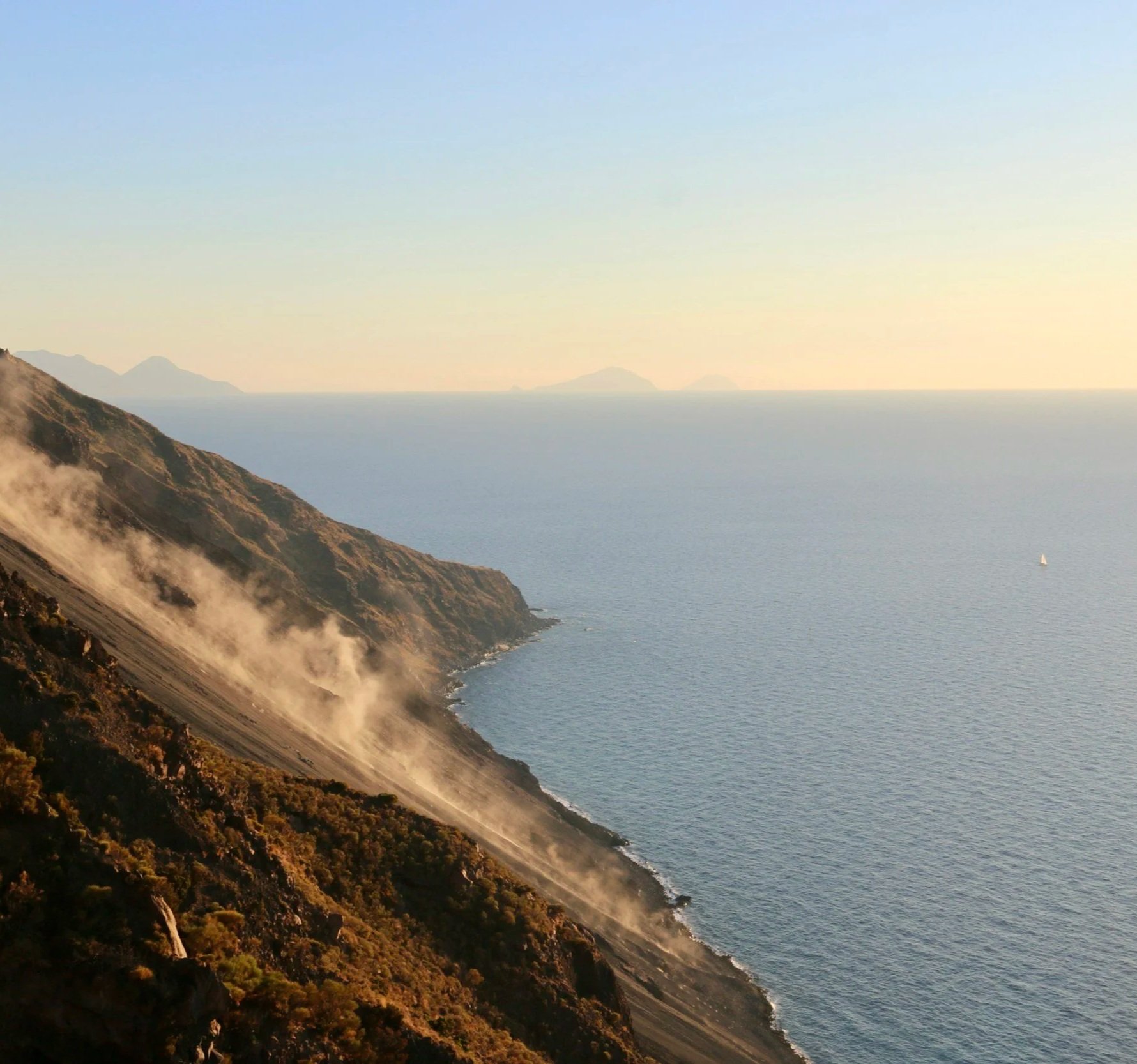 Coastal landscape with steep cliffs, fog or steam rising from the hillside, calm sea with a small sailboat, and distant islands under a clear sky.