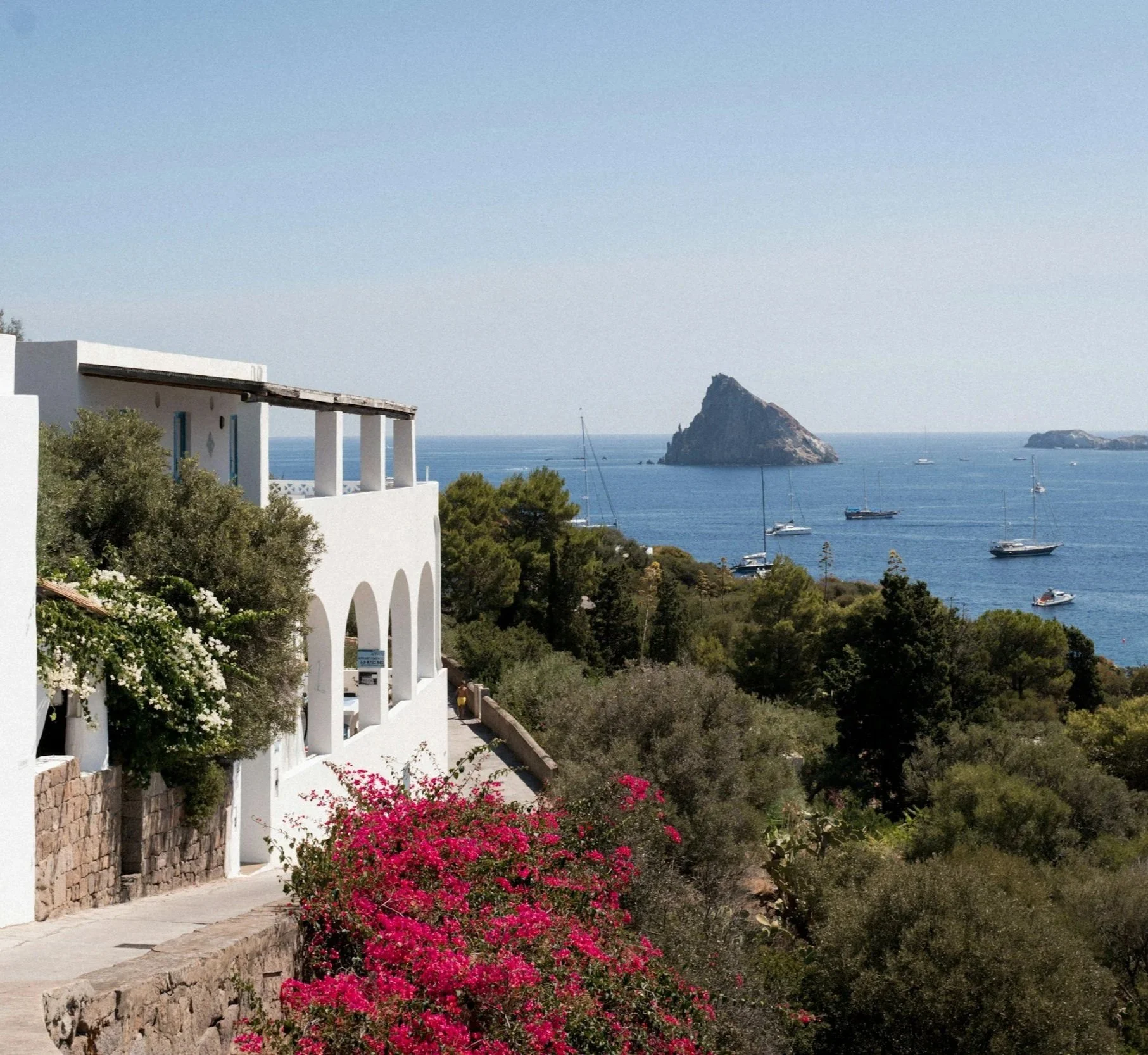 A scenic view of white Mediterranean buildings on a hillside with greenery, overlooking the ocean with several sailboats and small islands in the distance.