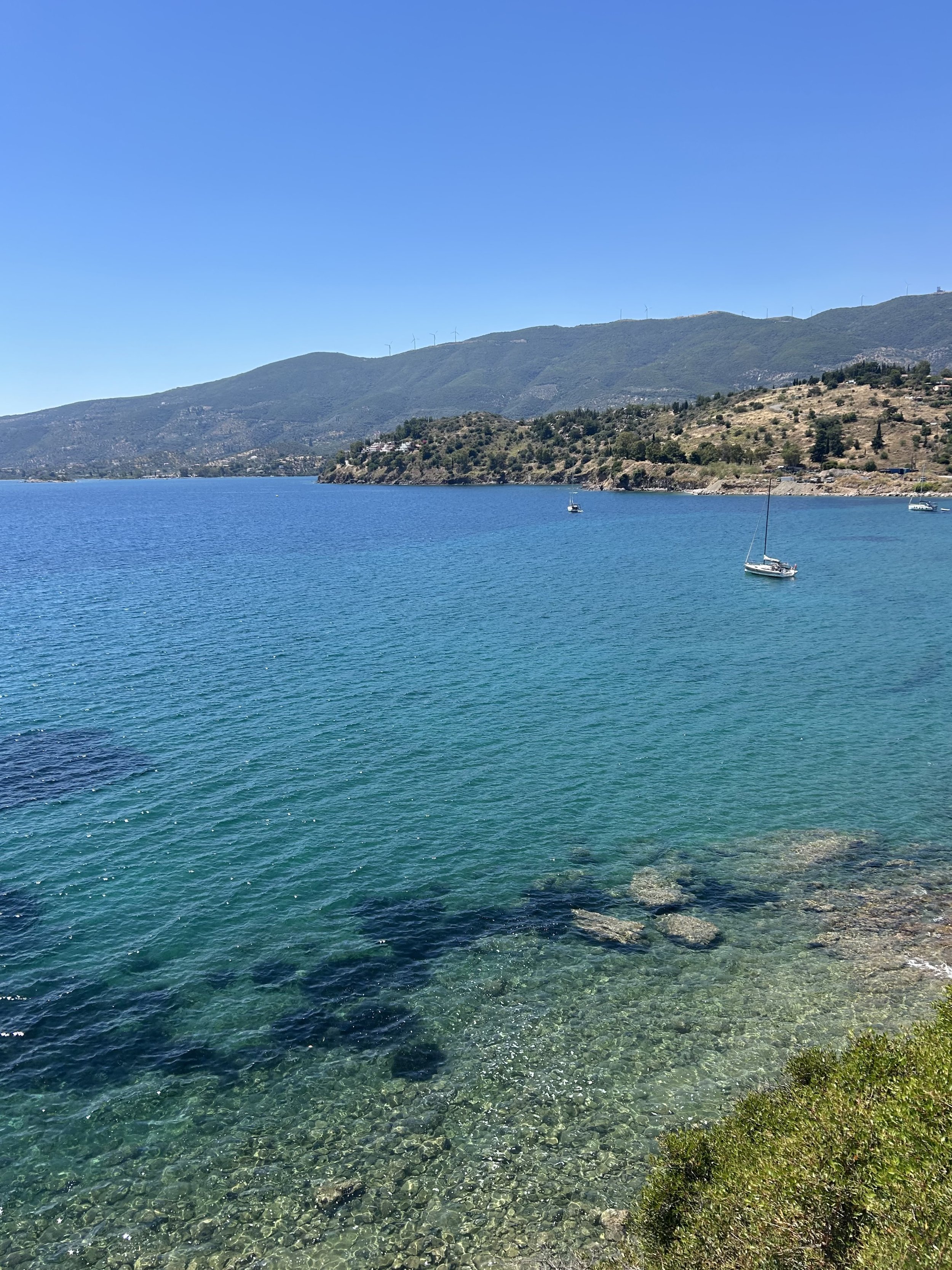 Clear blue water with a small sailboat and a mountain range in the background under a bright sky.