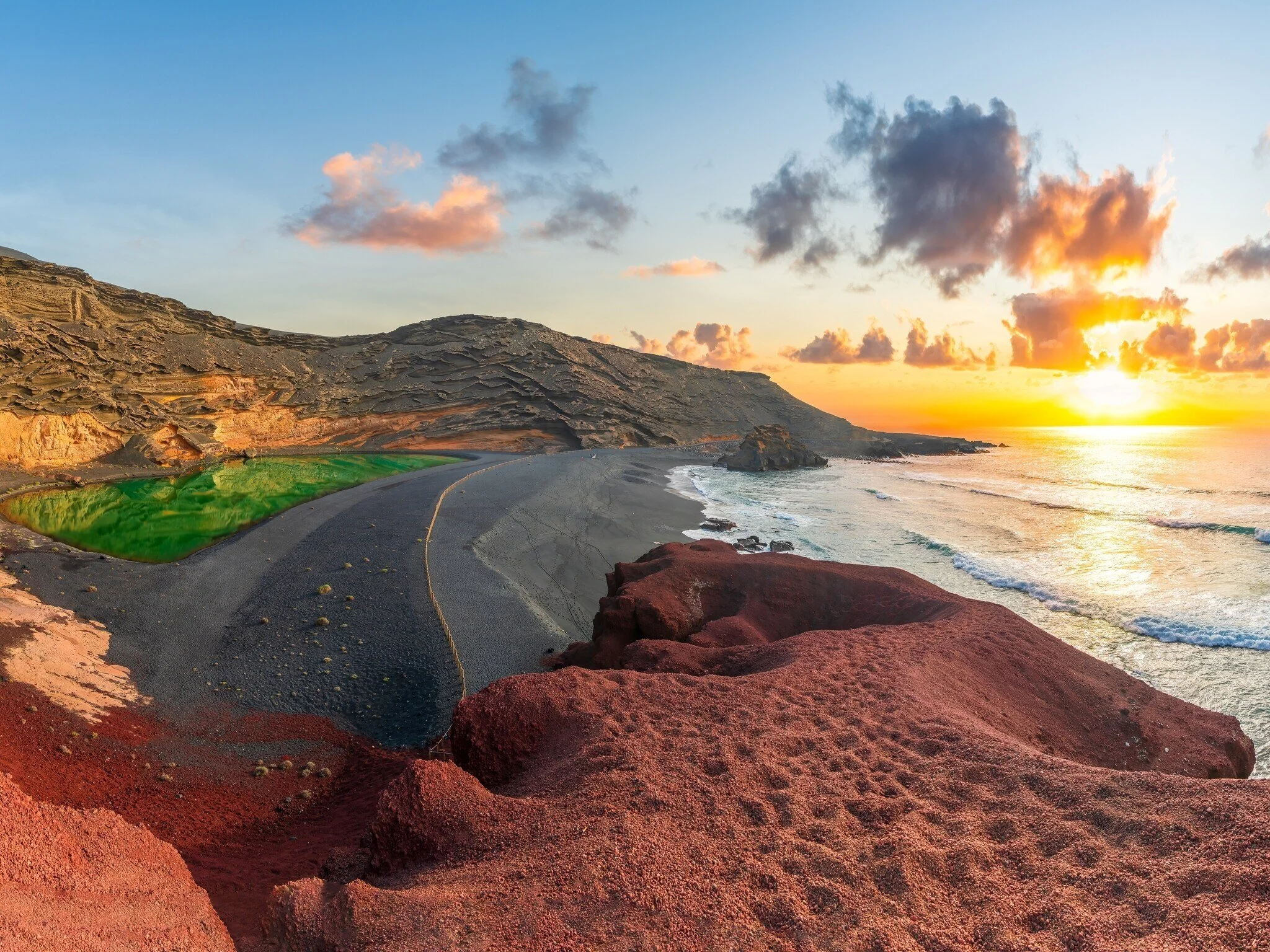 A coastal scene at sunset with a winding road along the rocky shoreline, cliffs on one side, and the ocean on the other. The sky is filled with clouds illuminated by the setting sun.