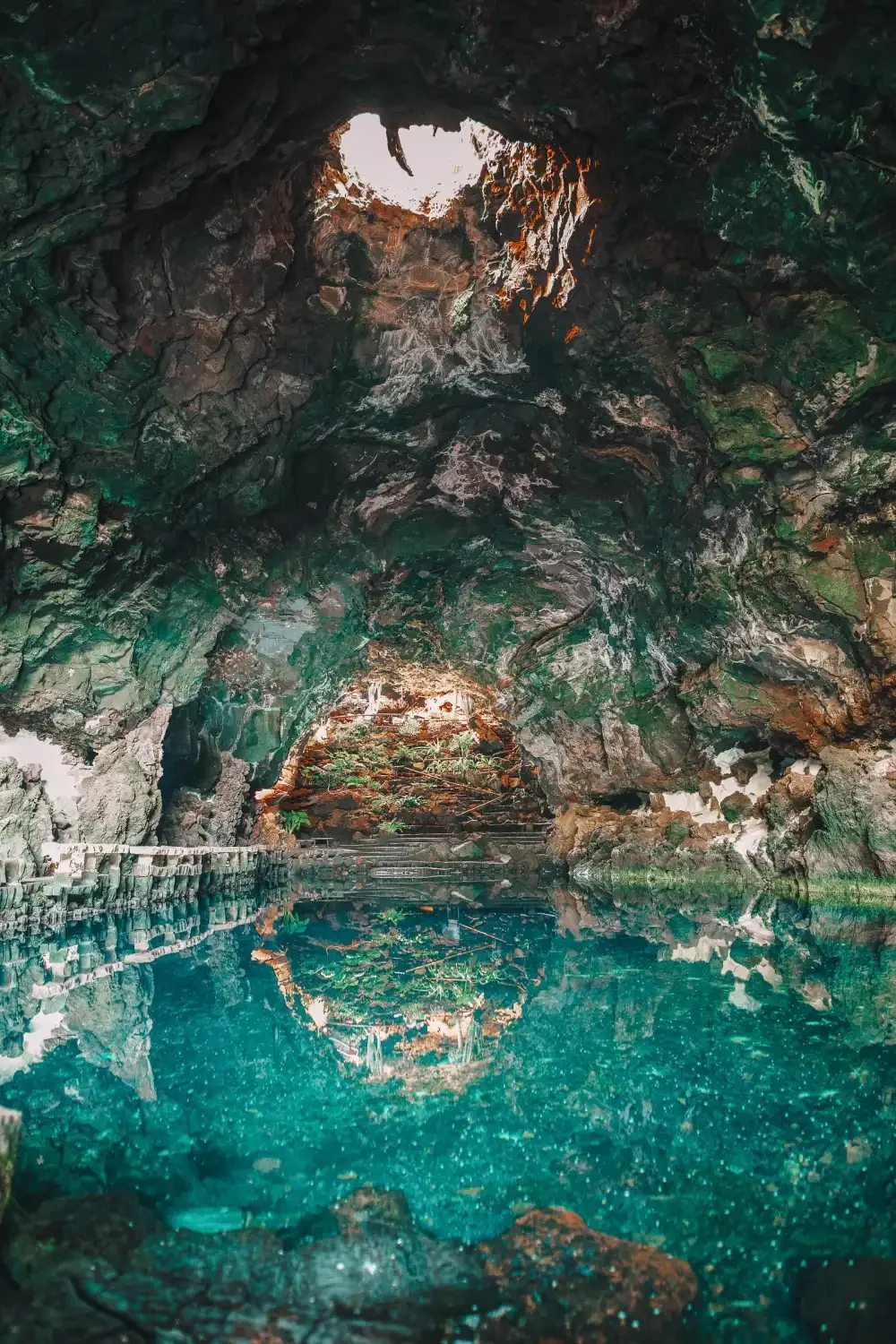 Inside a dark cave with a natural opening at the top, revealing a bright sky. The cave has rugged, textured walls with green and brown hues. A clear, turquoise pool of water reflects the cave's interior and the opening above.