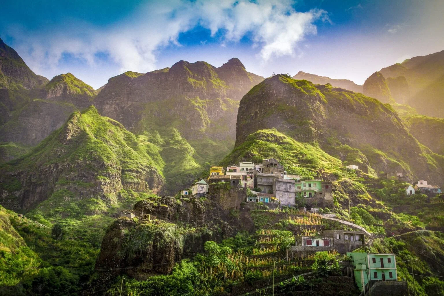 Scenic view of a small village built on a hillside surrounded by lush, green mountains with a partly cloudy sky.