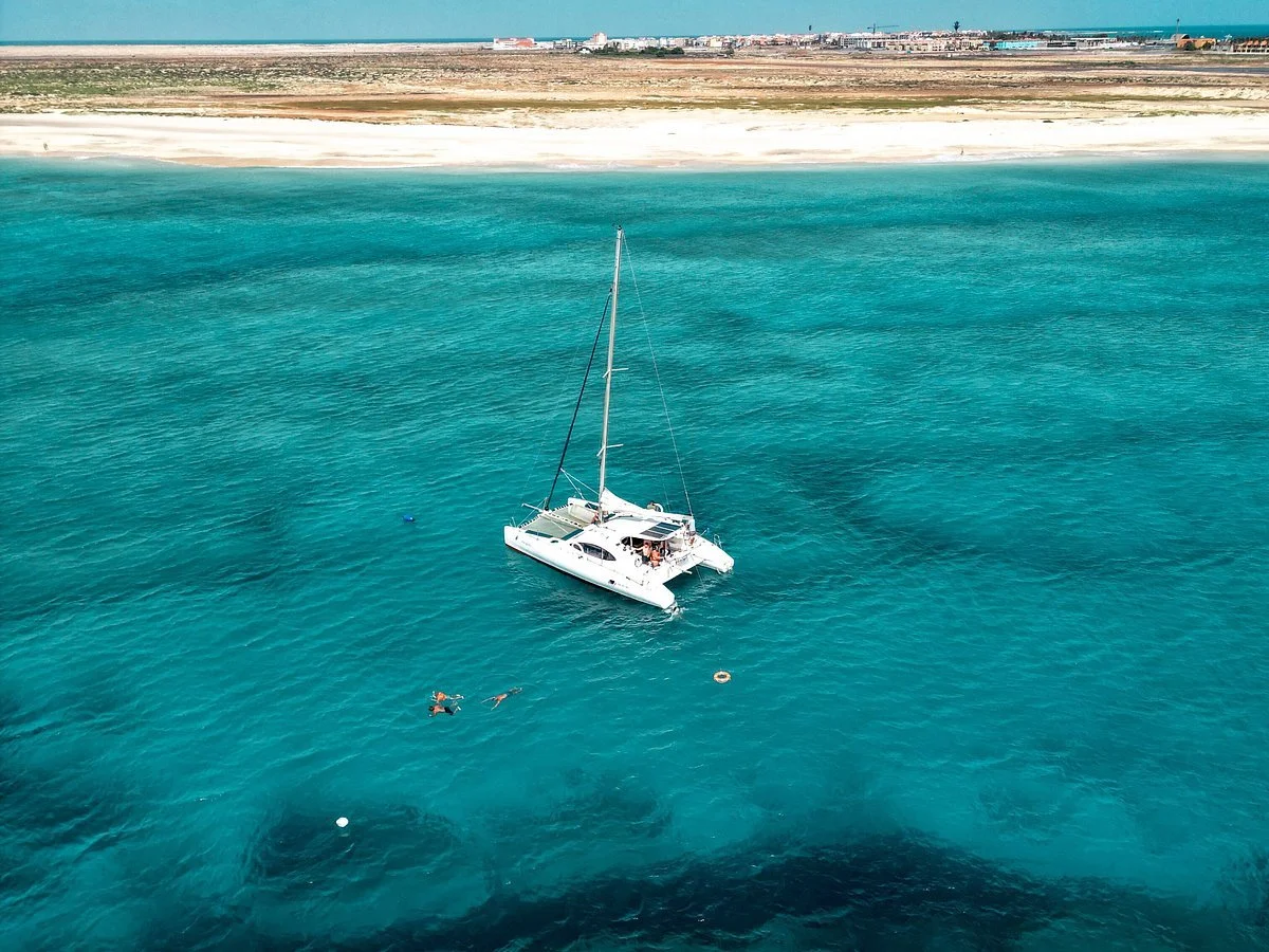 A white sailboat with people on board floating on clear blue ocean water near a sandy beach and distant shoreline.