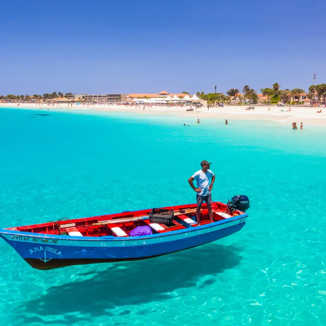 Man standing on a small blue boat in clear turquoise water with a beach and buildings in the background.