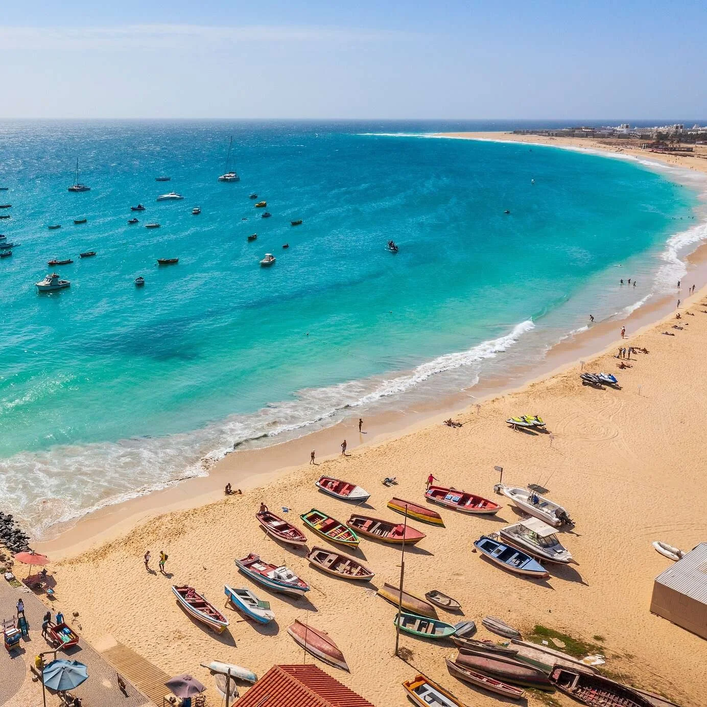 A beach scene with boats anchored in turquoise water, sandy shoreline, and a few people walking or sitting on the beach. Several boats are on the sand near a small building with a red roof.