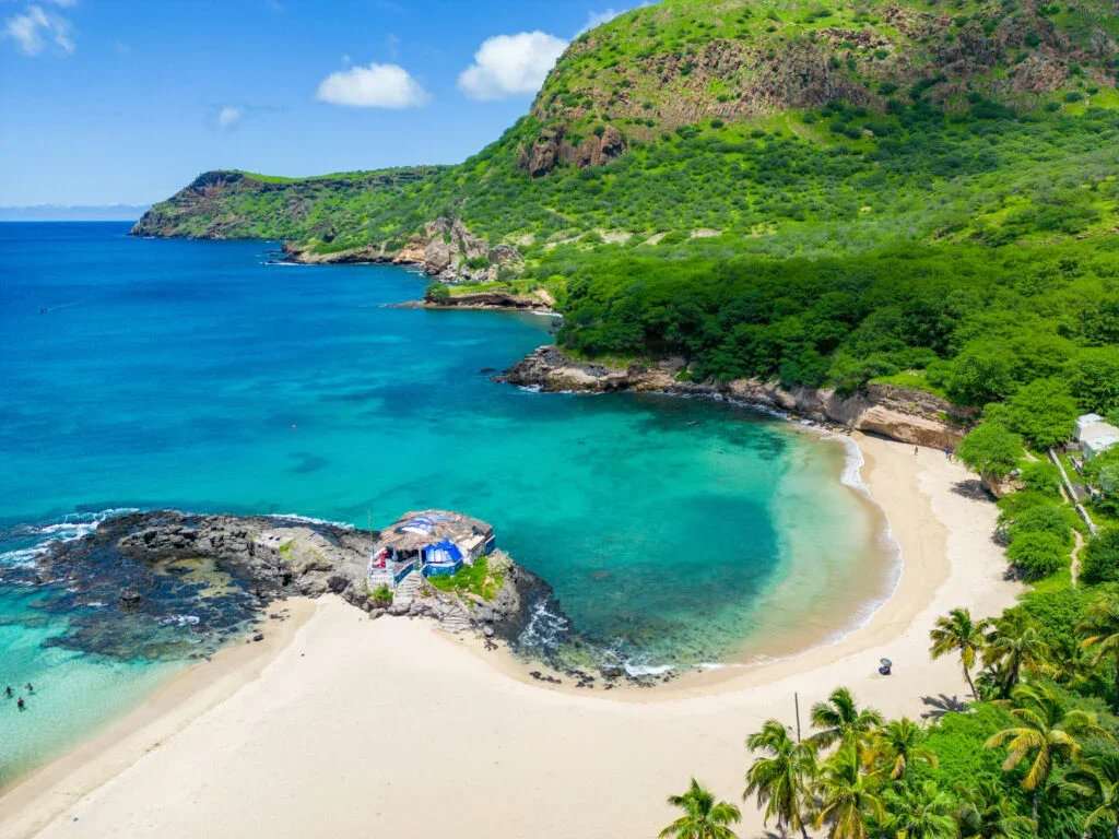 A tropical beach with white sand, turquoise water, lush green hills, palm trees, and a small rocky outcrop with umbrellas and chairs.