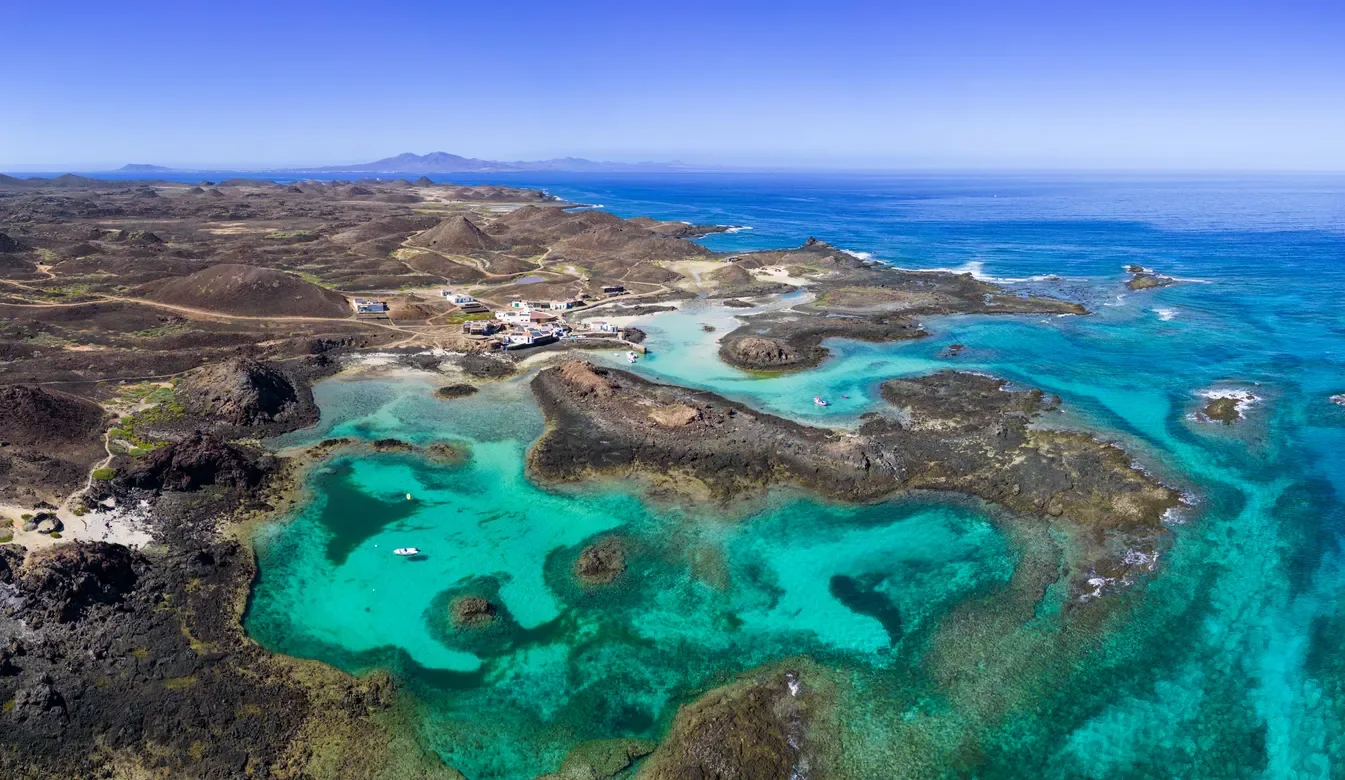 Aerial view of a coastal landscape with rocky terrain, turquoise waters, and small structures near the shoreline, with the open ocean in the background under a clear blue sky.