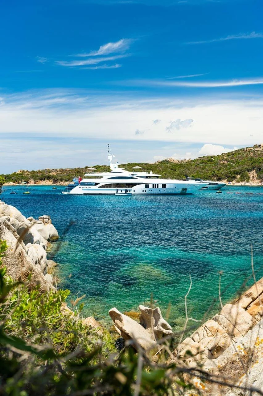 A luxury yacht floating on clear blue waters near a rocky and grassy shoreline under a partly cloudy sky.