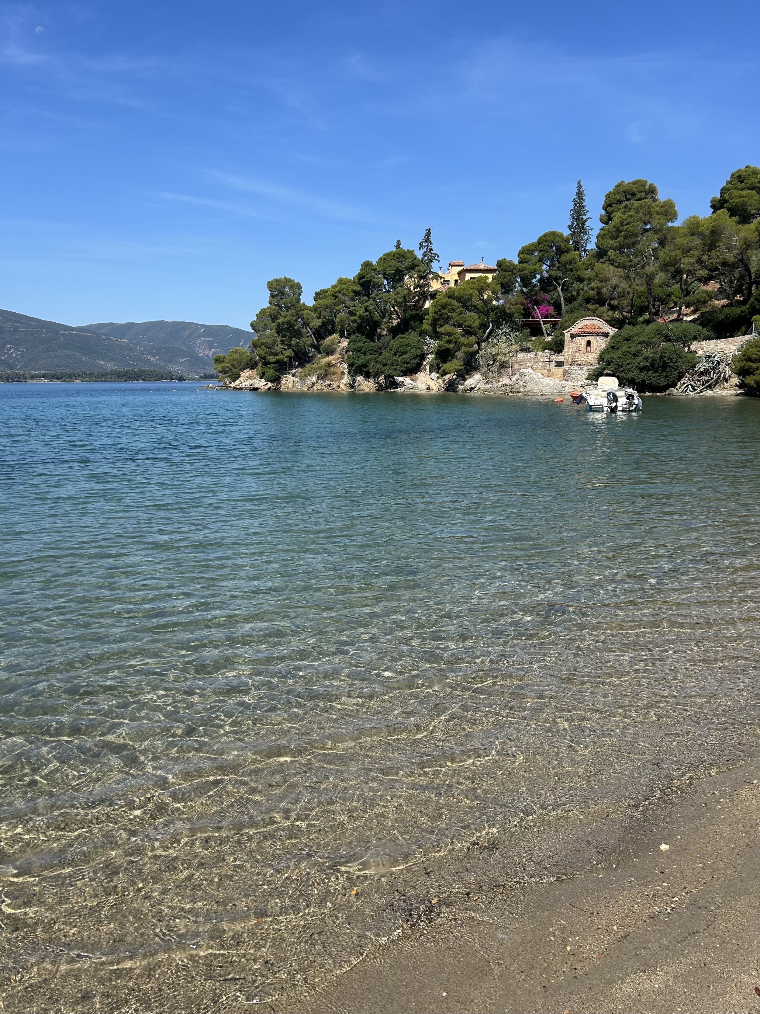 Clear water near a sandy shoreline with a lush green hillside in the background, and small buildings among trees, under a bright blue sky.