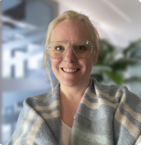 A woman with blonde hair wearing large round glasses, smiling, in an indoor setting with blurred background and plants.