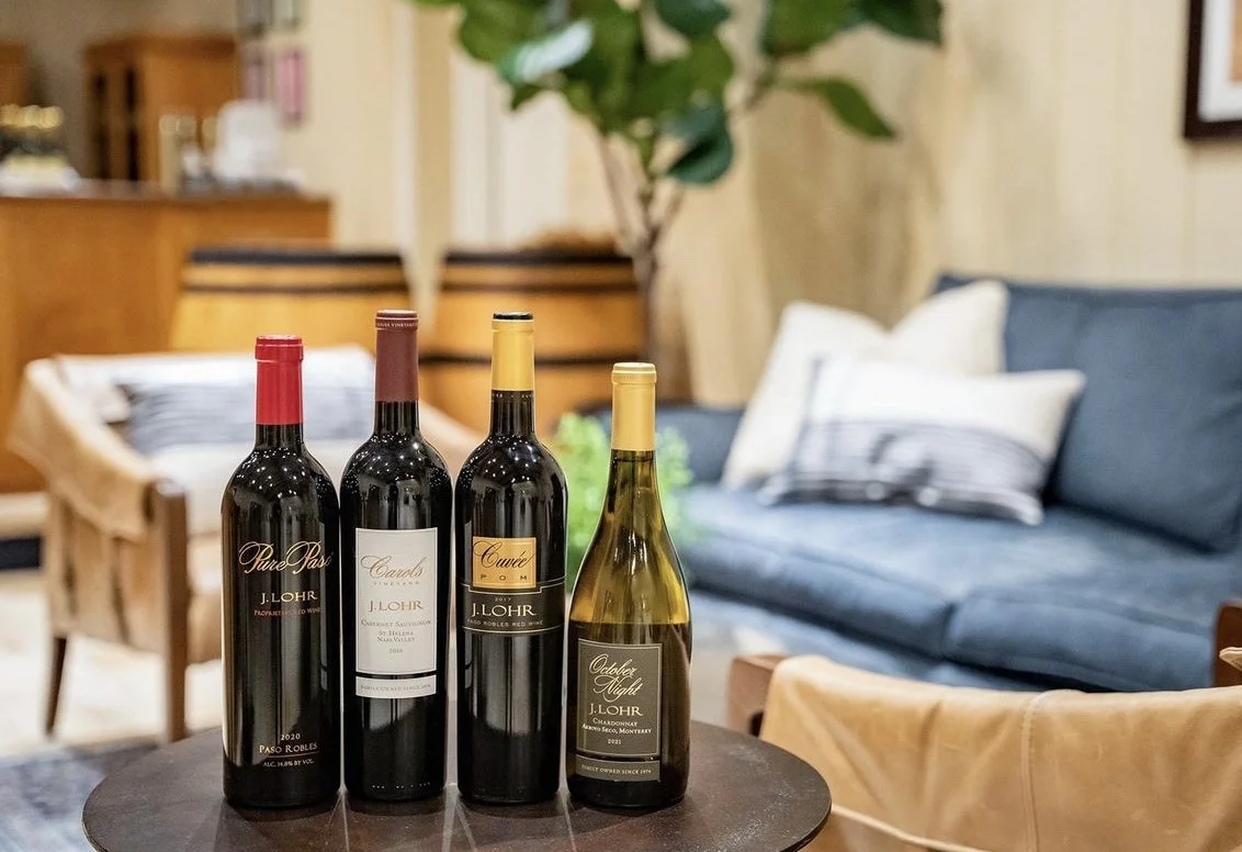 Four bottles of red wine on a round table in a cozy living room with a blue sofa, striped pillows, a wooden chair, and a potted plant in the background.