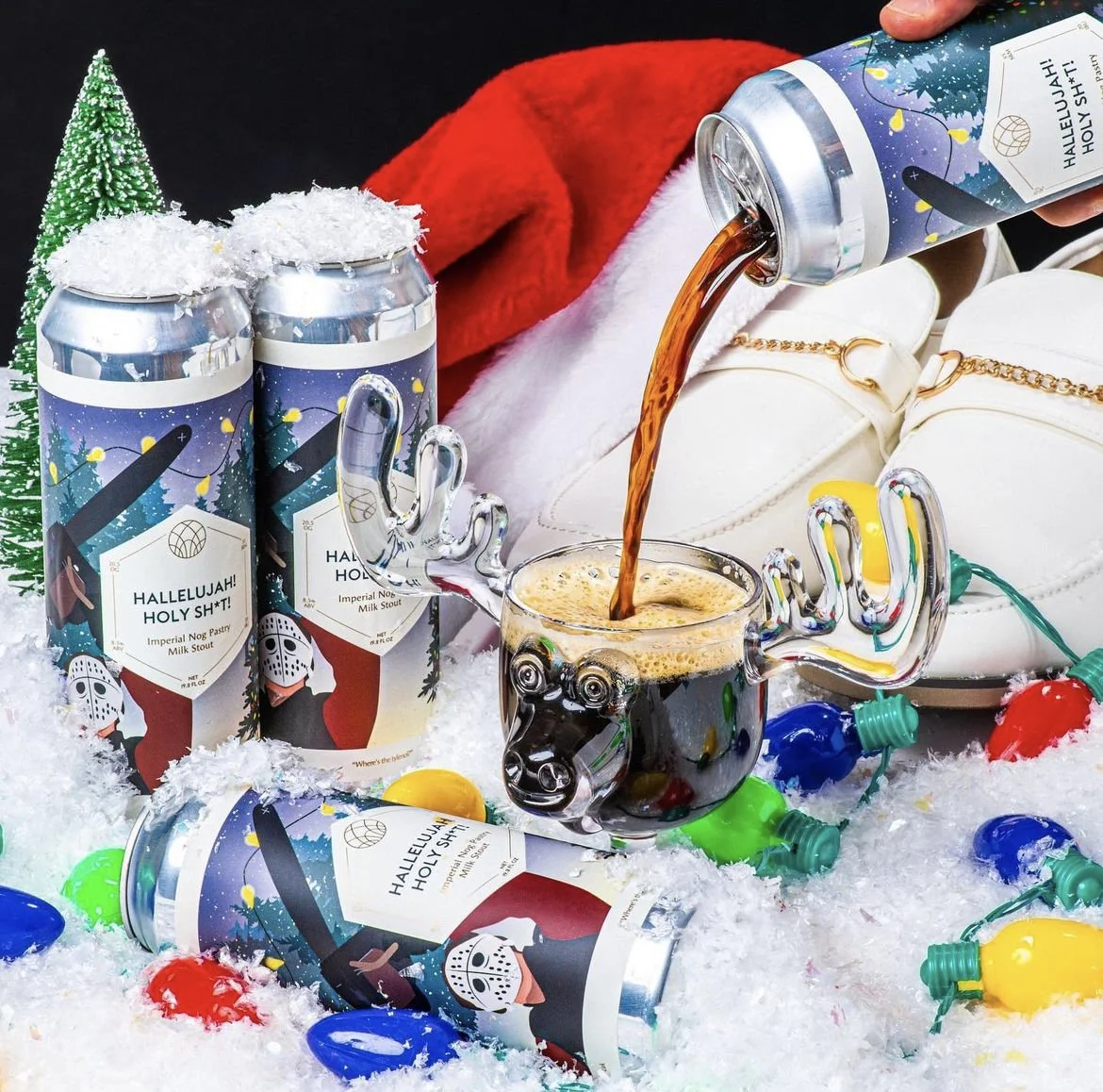 A festive scene with canned milk stout drinks labeled 'Hallelujah! Holy Sh*t!' surrounded by artificial snow, colorful Christmas lights, a green Christmas tree, a red and white Santa hat, white shoes, and a glass mug shaped like a pig's head filled with dark beer.