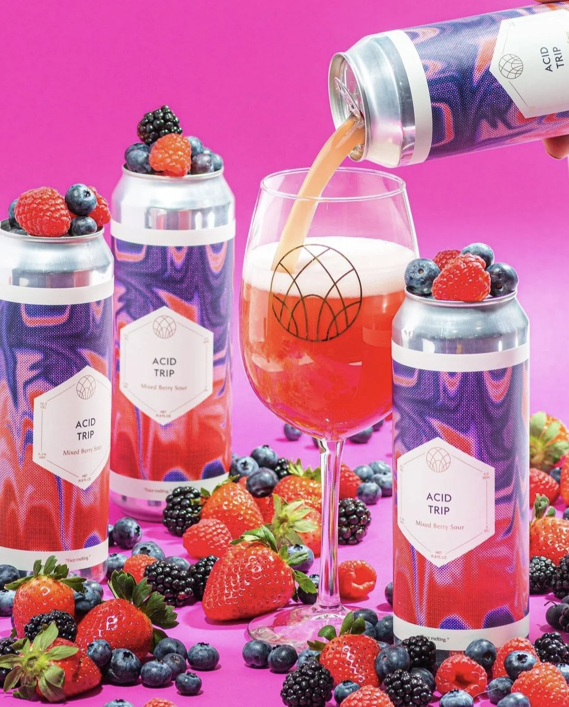 Cans of mixed berry flavored drink labeled 'Acid Trip,' with a glass of pink berry concoction being poured and surrounded by fresh strawberries, blackberries, and blueberries on a pink background.
