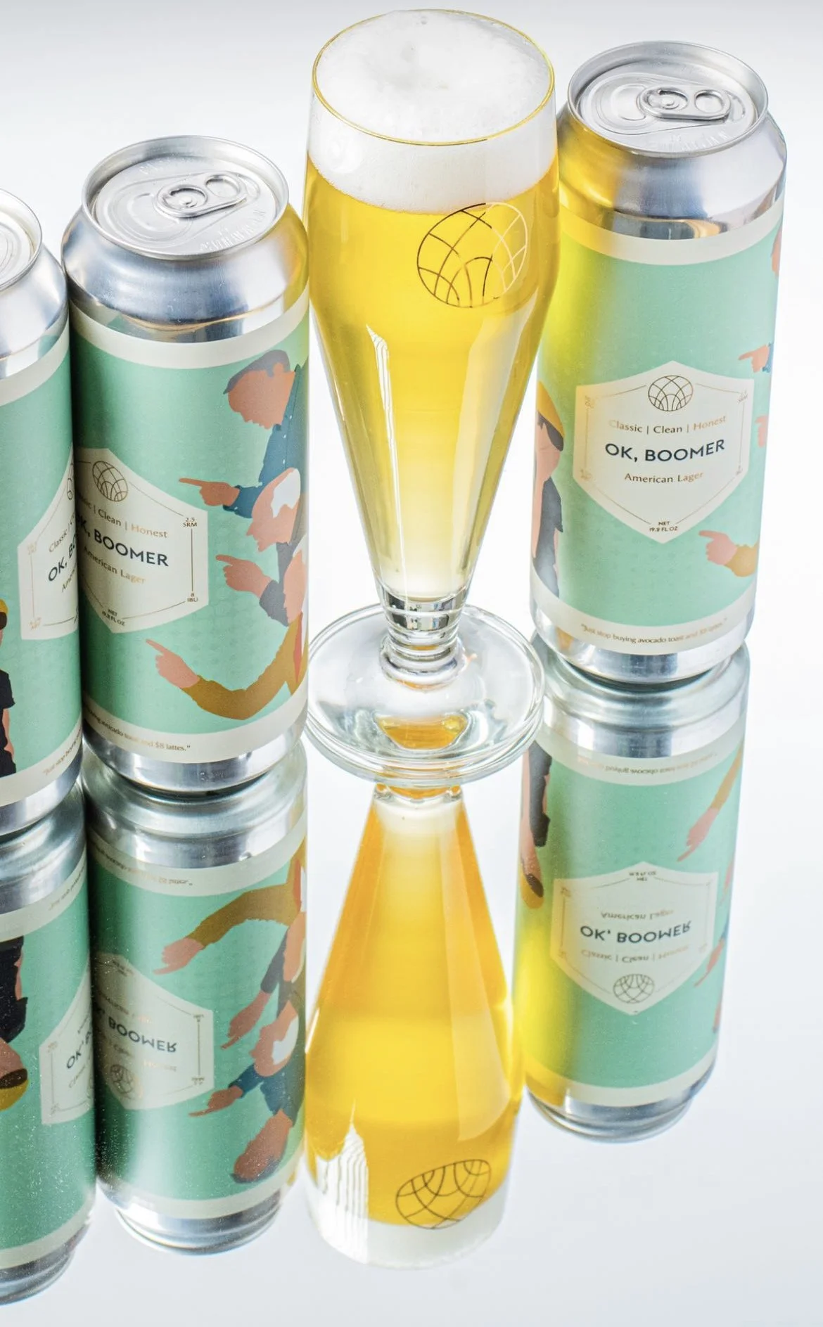 Cans of OK, BOOMER American lager beer and a glass of beer with foam, arranged on a reflective surface.