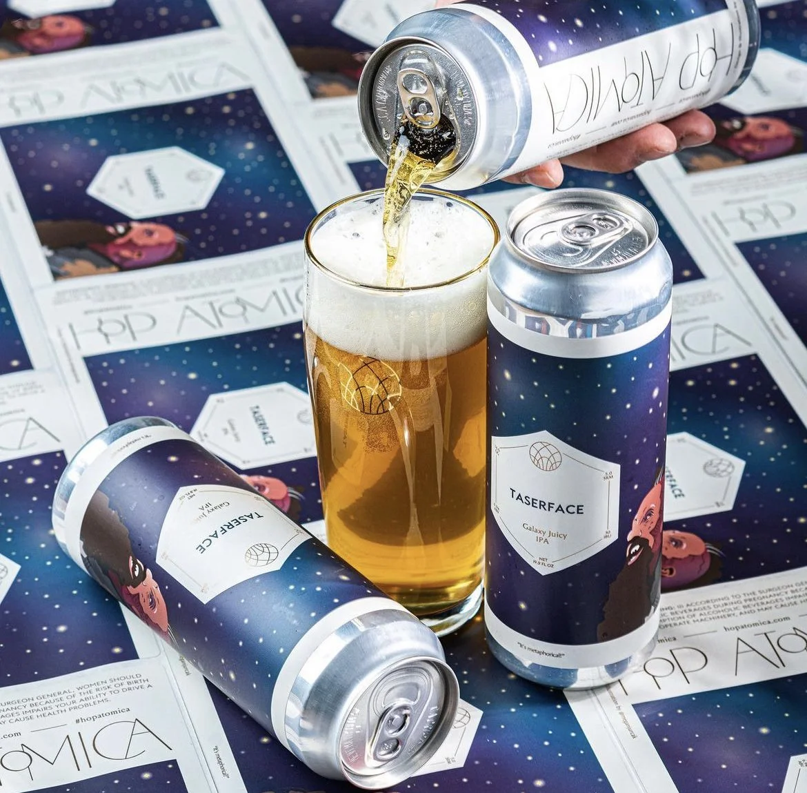 A can of Taserace Galaxy Juicy IPA being poured into a glass, with two additional cans on a galaxy-themed table surrounded by flyers or posters featuring a cosmic design and a cartoon character.