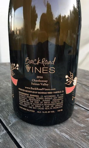 Bottle of Back Road Vines 2016 Chardonnay wine with a black label and gold decorative elements, reflecting outdoor scenery.