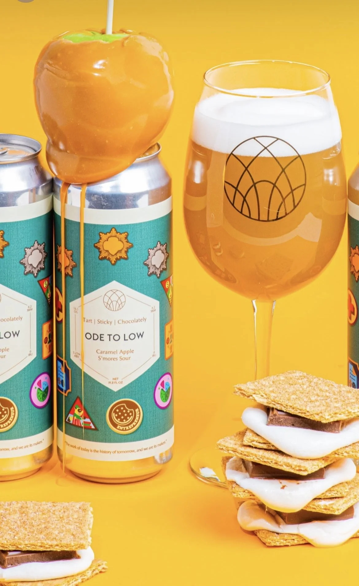 A caramel apple with green and orange icing on a stick, a glass of beer, a stack of caramel apple s'mores with graham crackers, marshmallow, and chocolate, and cans of craft beverage, all against a bright yellow background.