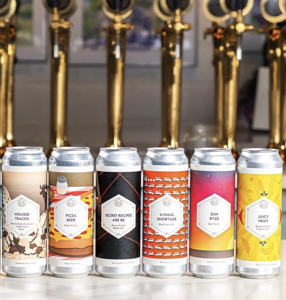 Six colorful cans of craft beer with unique labels are displayed in front of a row of gold-colored taps.