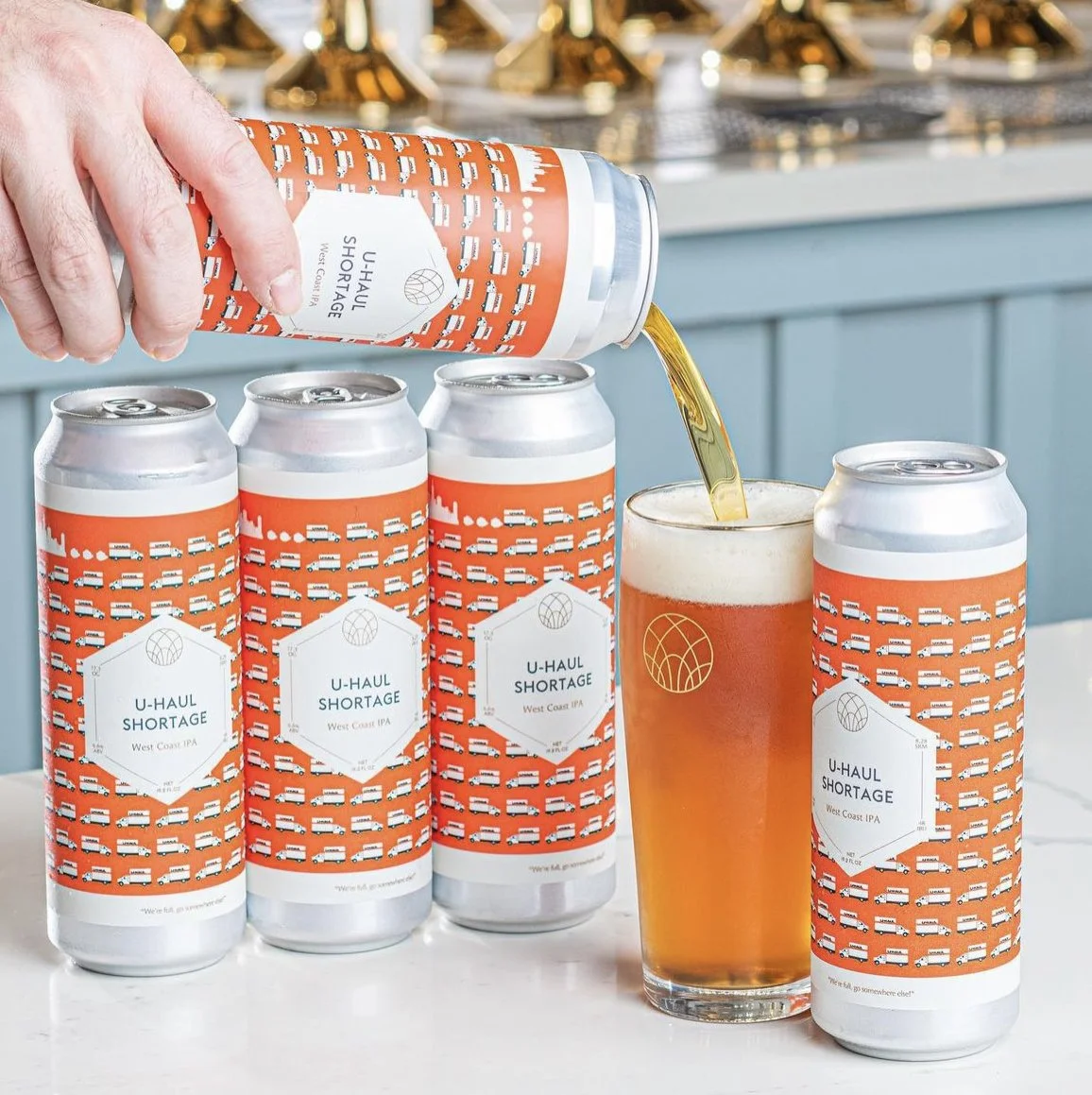 Pouring a craft beer from a can labeled 'U-Haul Shortage West Coast IPA' into a glass, with several similar cans in the background.