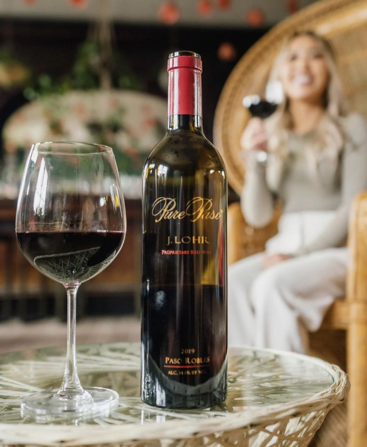 A bottle of red wine and a glass of red wine on a wicker table, with a woman holding a glass of wine in the background, sitting in a wicker chair.