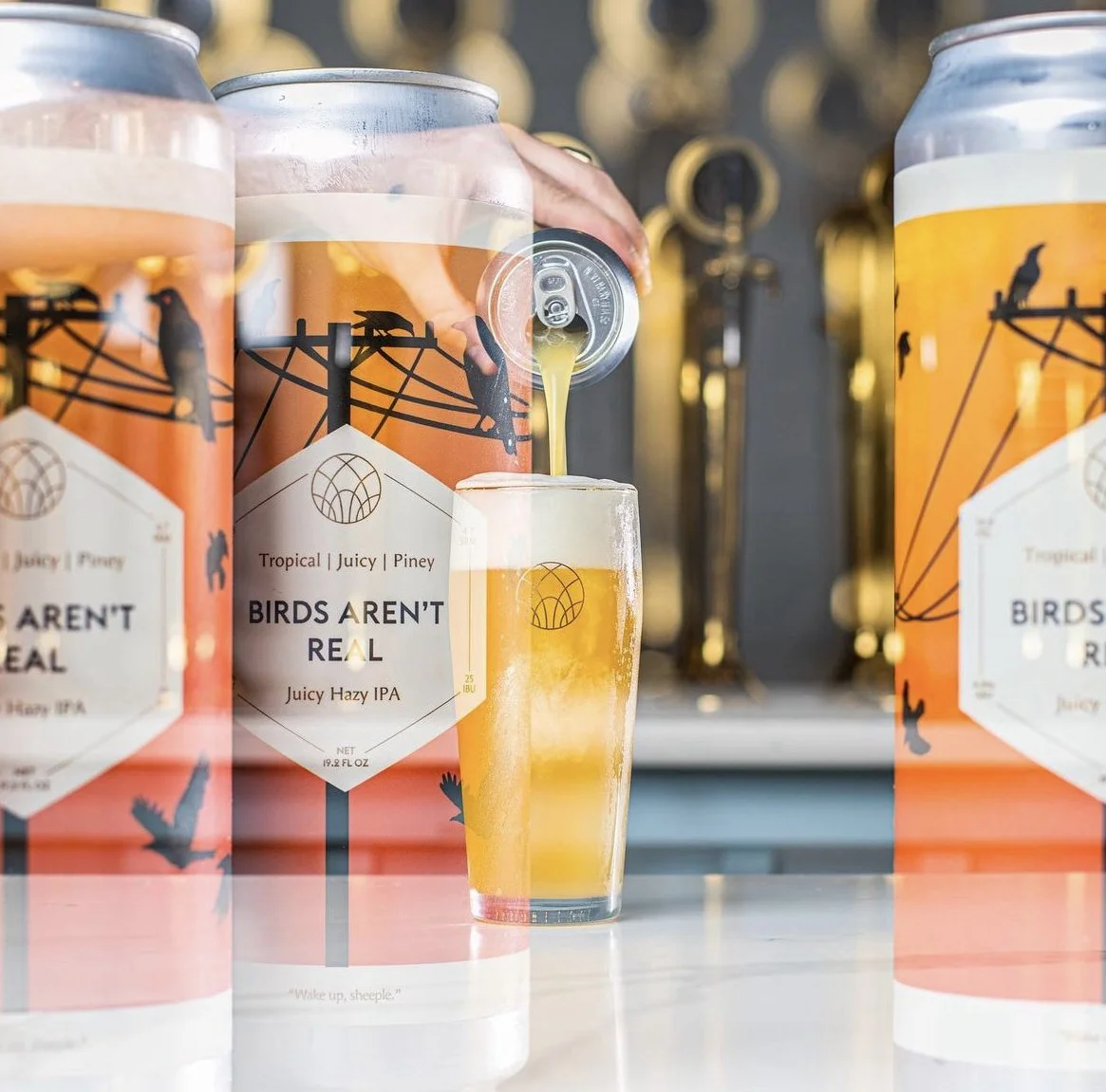 A can of Birds Aren't Real Juicy Hazy IPA beer being poured into a glass, with some cans visible in the foreground, against a blurred background of a wall with hooks and hanging tools.