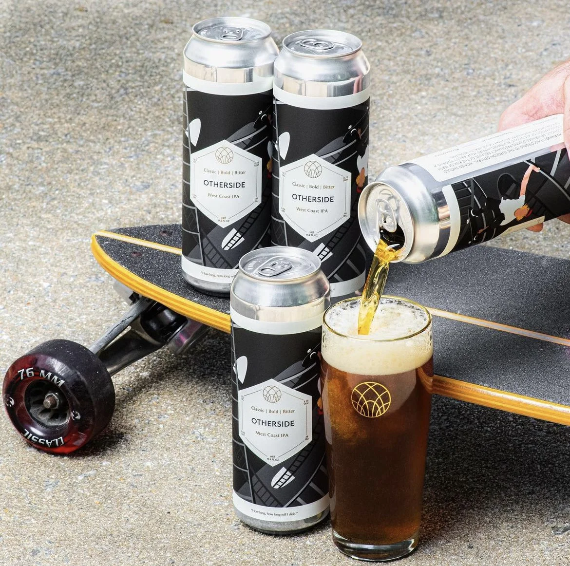 A skateboard on a sandy surface with four cans of 'Ourside' West Coast IPA beer, one can being poured into a glass. The skateboard has a black deck with a yellow edge. The beer in the glass is amber with a foamy head.