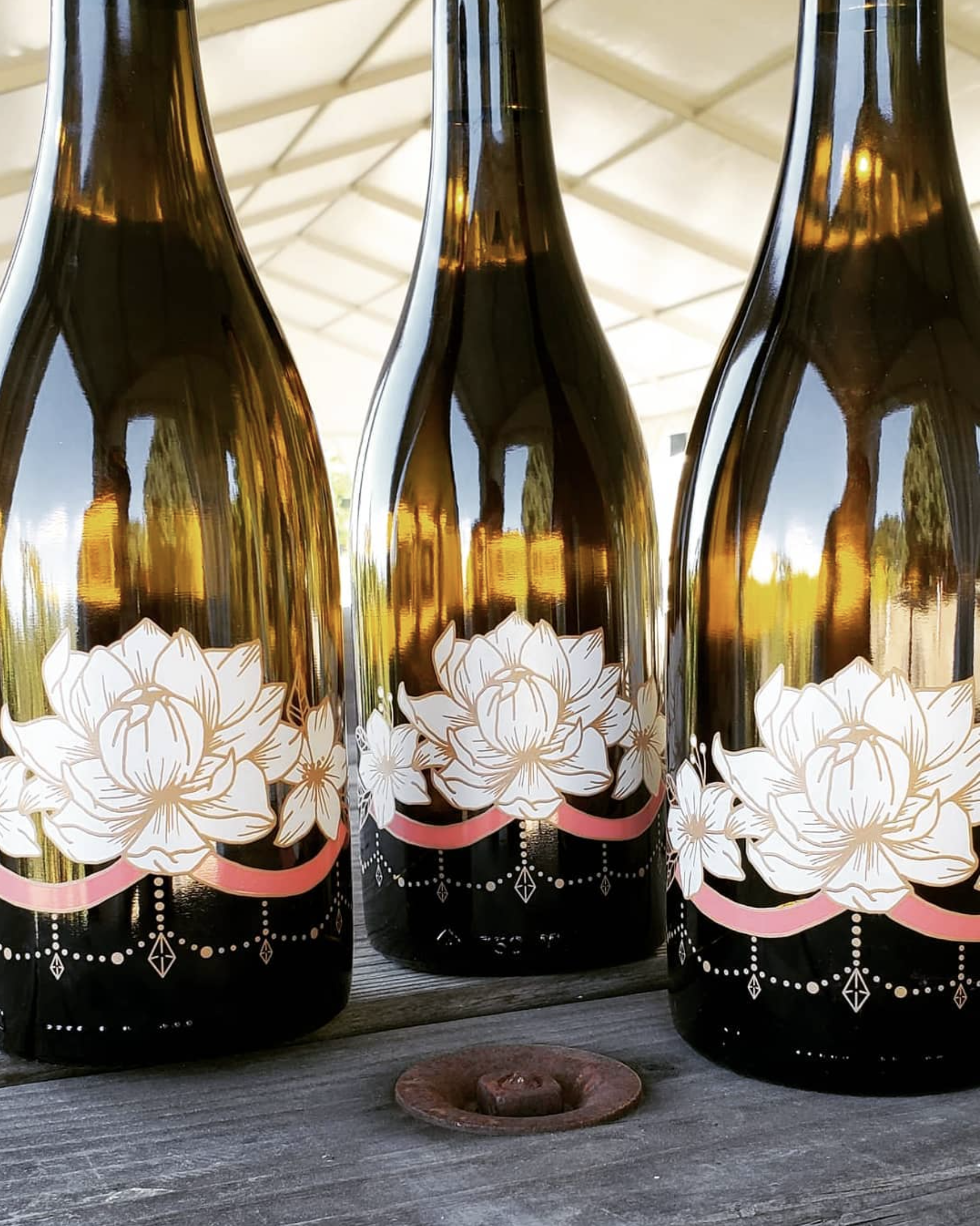 Three amber glass bottles with white floral designs and pink accents, placed on a wooden surface outdoors.