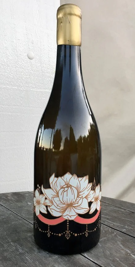 A black bottle with a gold cap and a floral design in pink, gold, and white on the lower half, placed on a dark wooden surface.