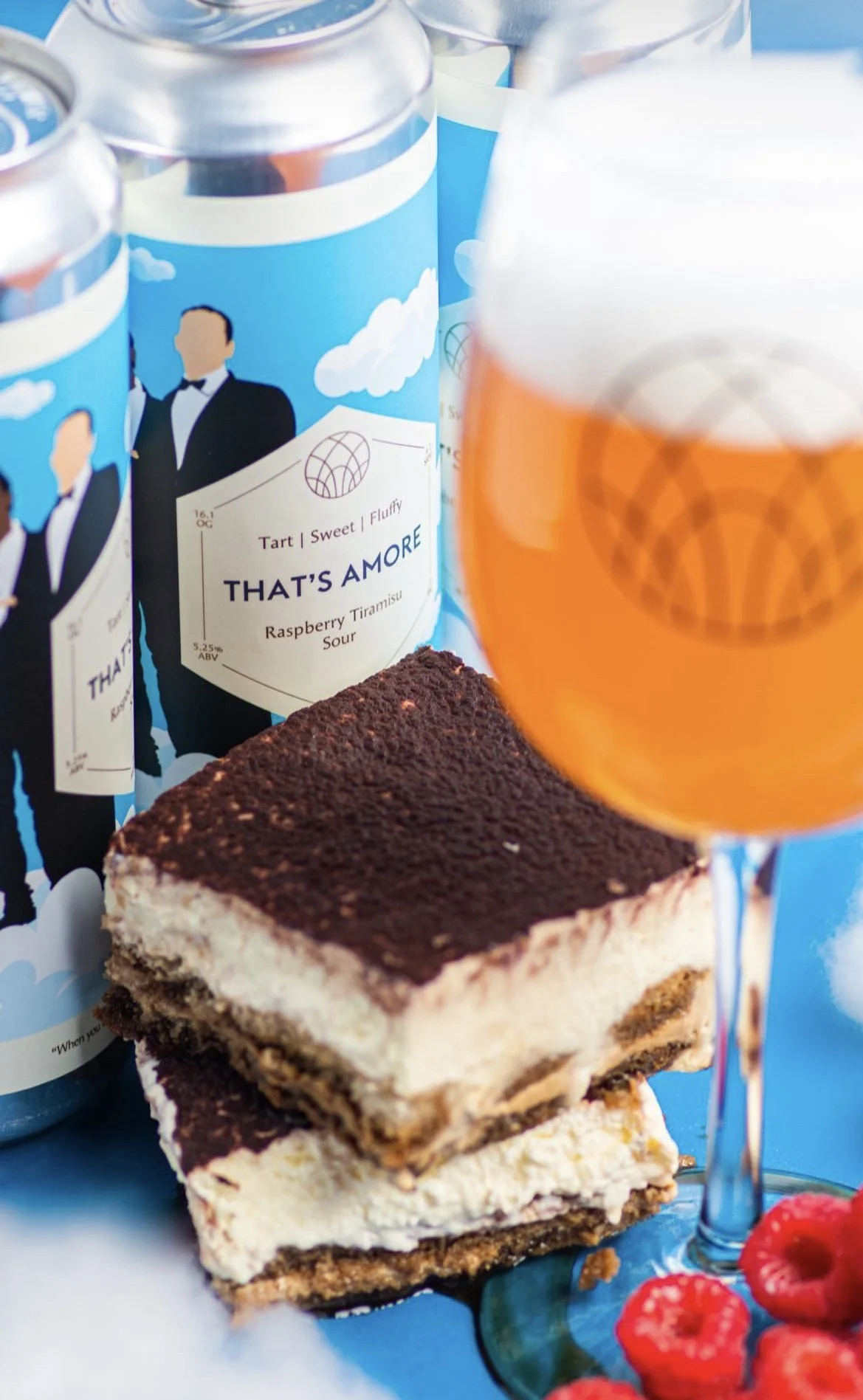 A close-up of a dessert bar with layers of cream, biscuit, and chocolate, several raspberry fruits, a glass of orange drink with foam, and cans of beverage with a label reading 'THAT'S AMORE Raspberry Tiramisu Sour' in the background.