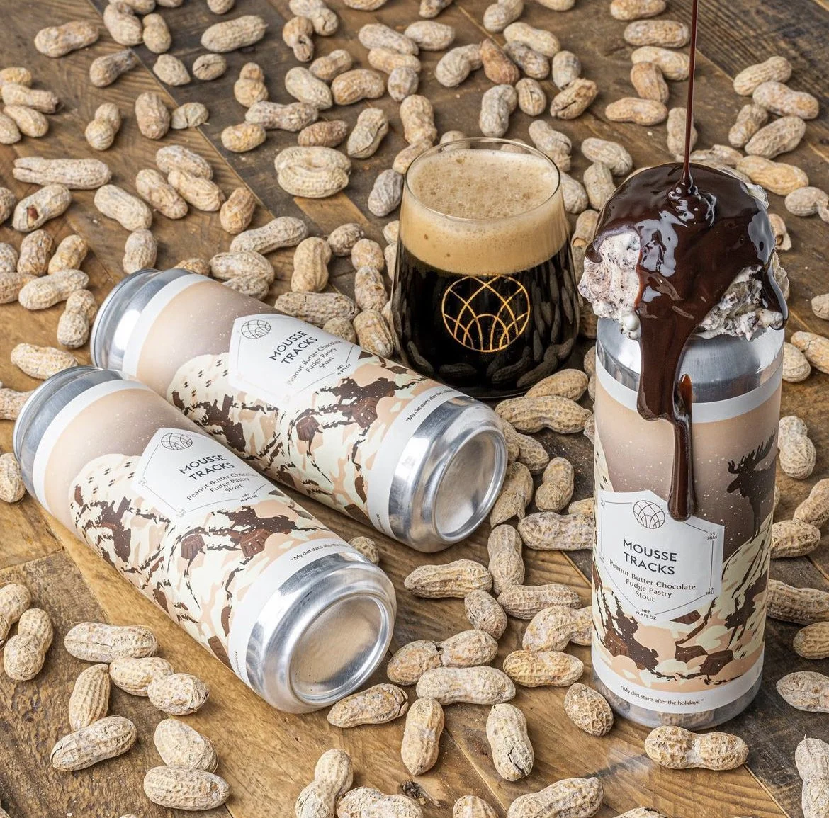 Cans of peanut butter chocolate mousse in a rustic setting with peanuts scattered around, a glass of dark stout beer, and a dessert topped with chocolate sauce on a wooden surface.