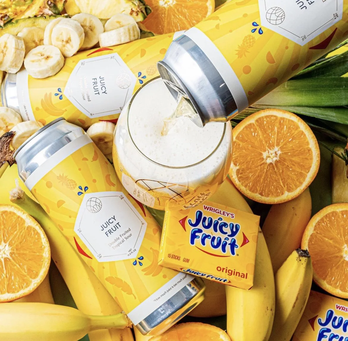 Can of Wrigley's Juicy Fruit gum being poured into a glass of juice surrounded by oranges, bananas, pineapple, and Juicy Fruit gum packages.