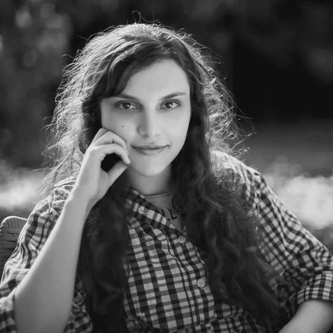A photo of Mahtab Rezayi, an Iranian filmmaker based in Austin, TX.  Black and white portrait of a young woman with long wavy hair, wearing a checkered shirt, sitting outdoors with a blurred background, looking at the camera with a slight smile.