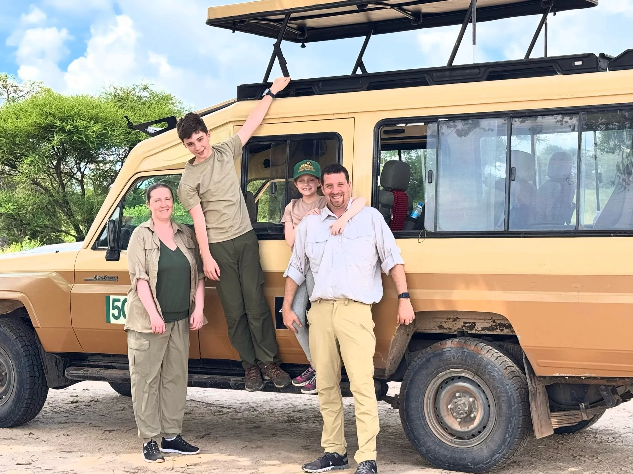 Group of four people, two children and two adults, standing in front of a safari vehicle outdoors with trees and blue sky in the background. The children are on the vehicle's door, and the adults are standing on the ground.