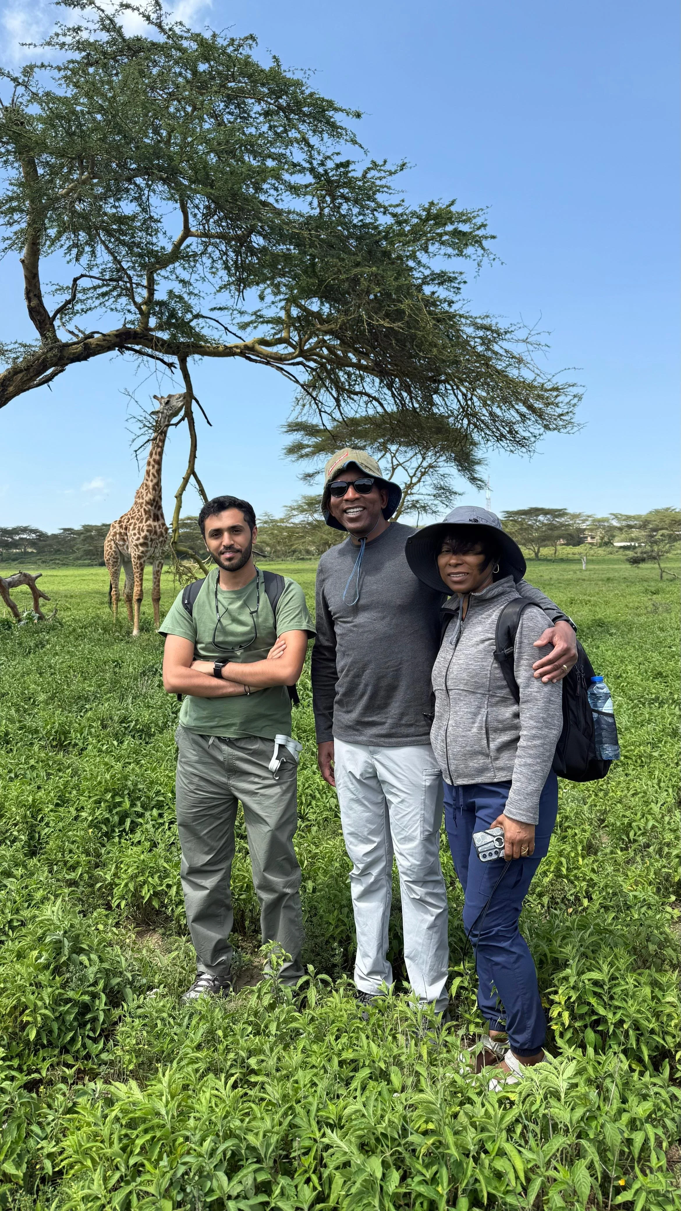 Three people standing together in a lush green field with a giraffe and a tree in the background, under a clear blue sky.