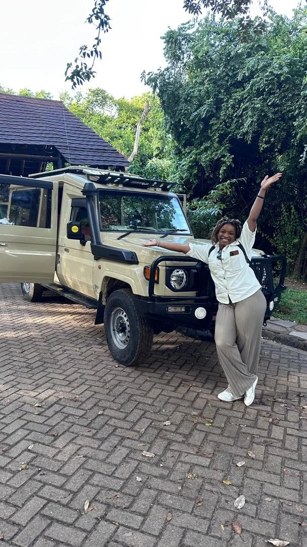 A woman standing next to a beige off-road vehicle with the door open, smiling and gesturing with her arms, in an outdoor setting with trees and a paved ground.