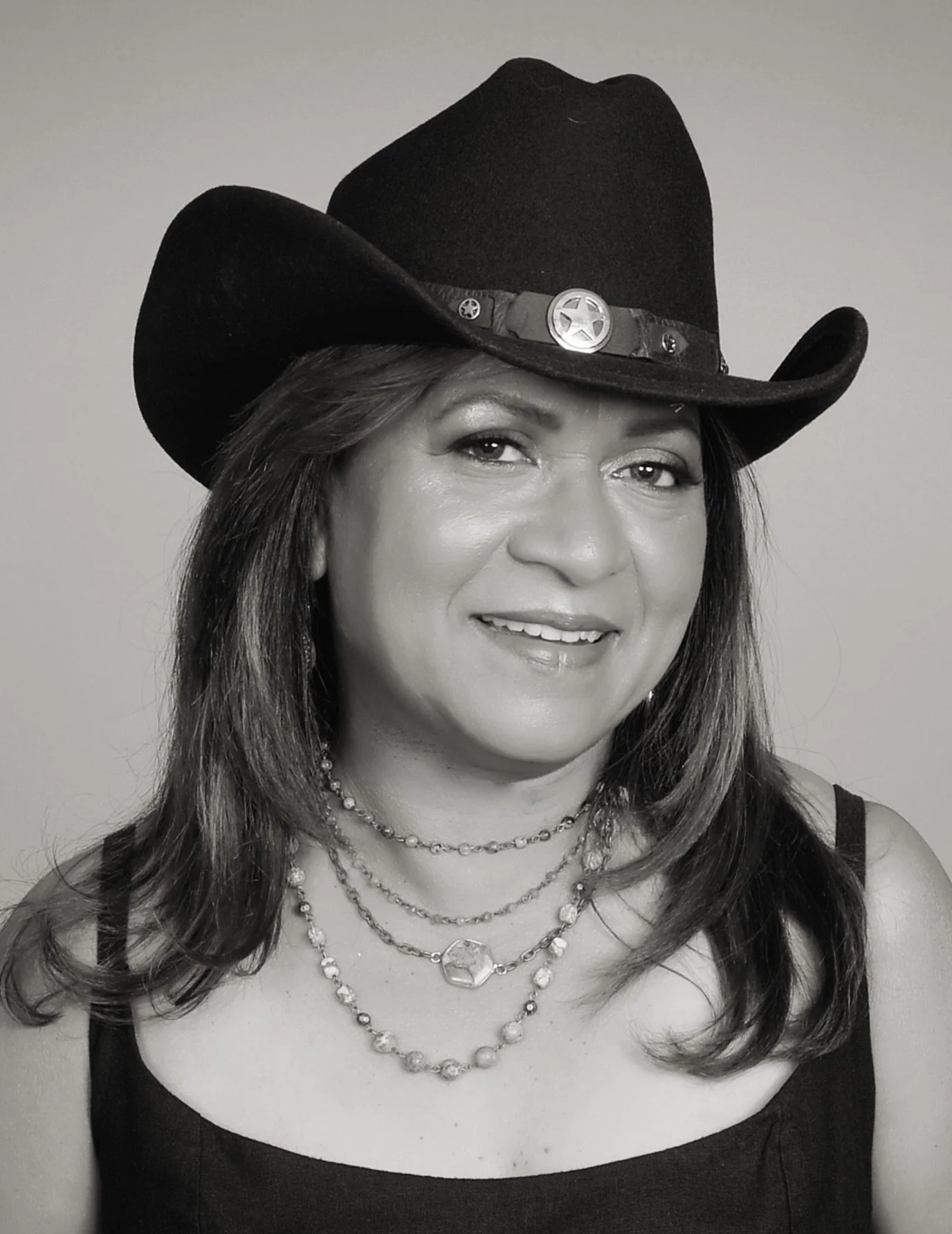 A woman wearing a cowboy hat, layered necklaces, and a tank top, smiling at the camera.