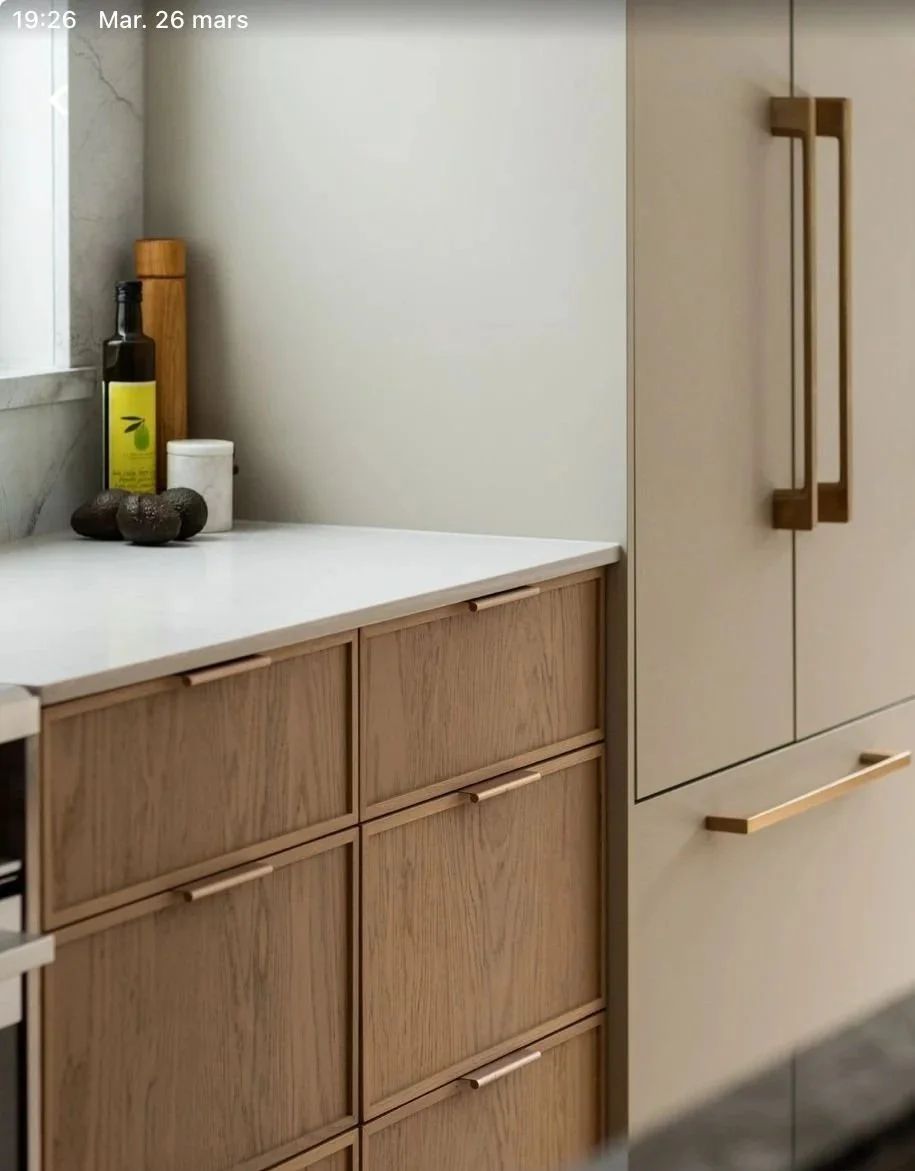 Close-up of a modern kitchen counter with wooden cabinets, a dark bottle of olive oil, a white container, and some decorative stones near a window.