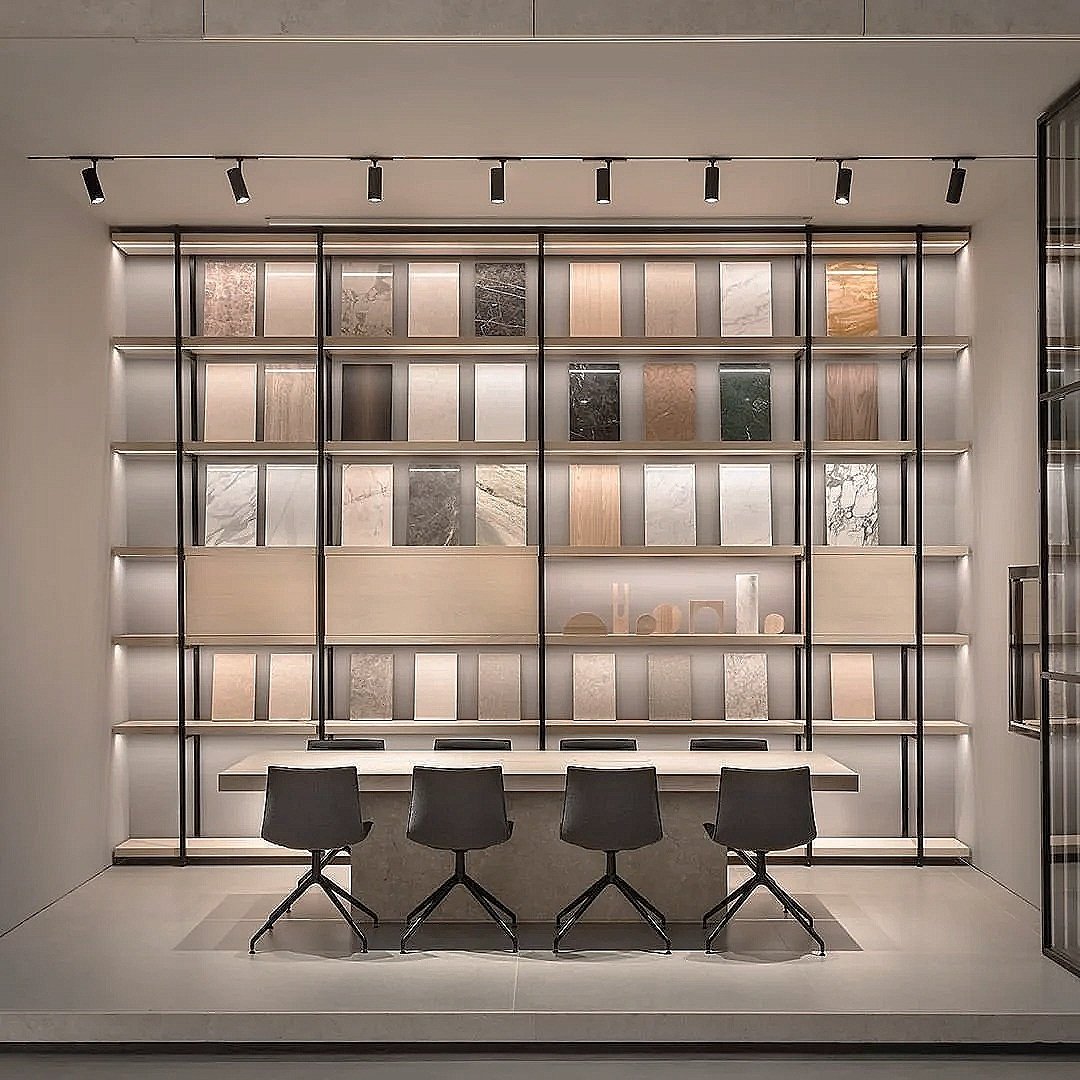 Modern meeting room with four black chairs at a white table, in front of a wall display featuring various tiles and materials, illuminated by ceiling spotlights.