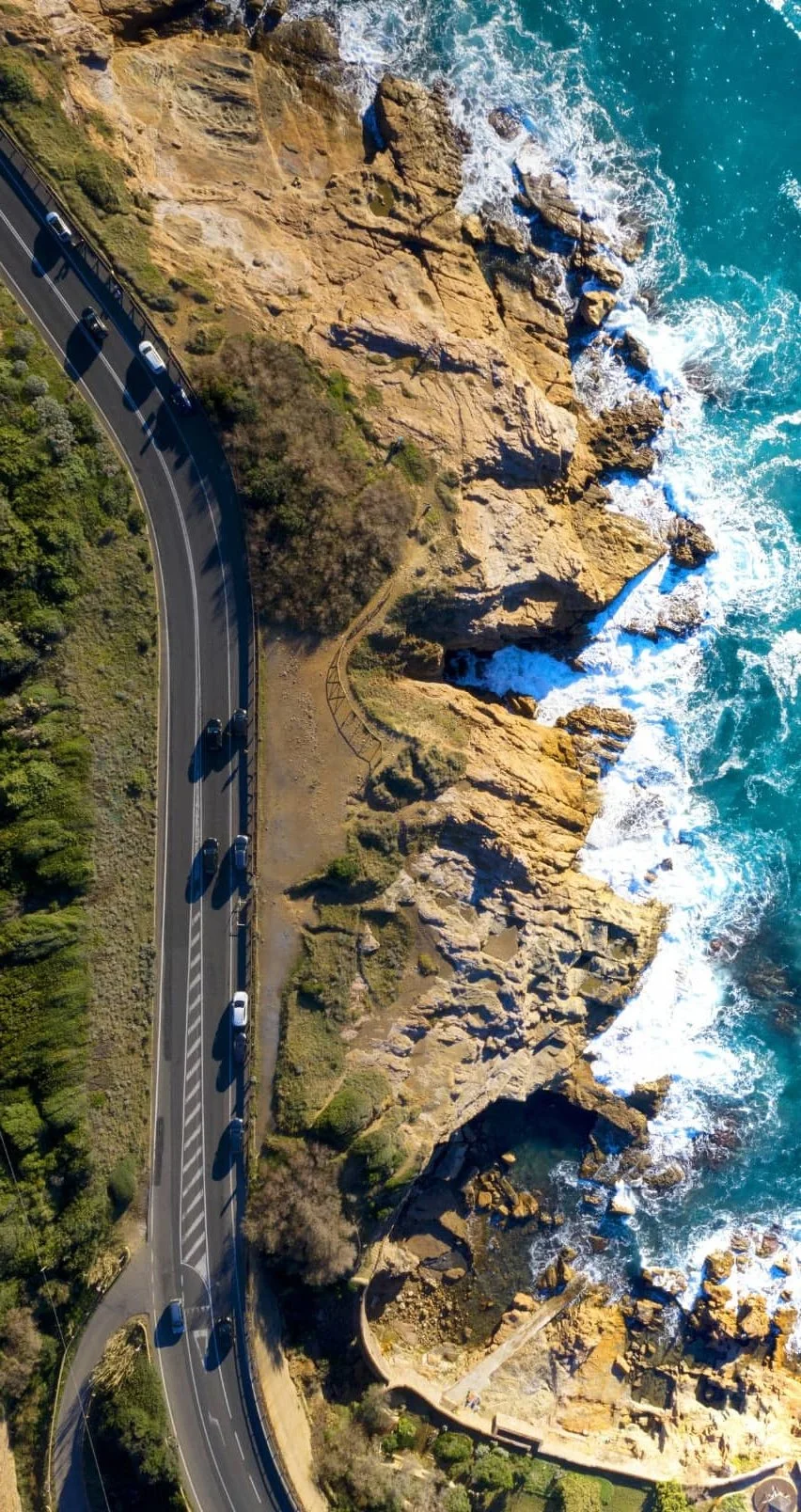 Aerial view of a winding coastal road running parallel to rocky shoreline and ocean waves.