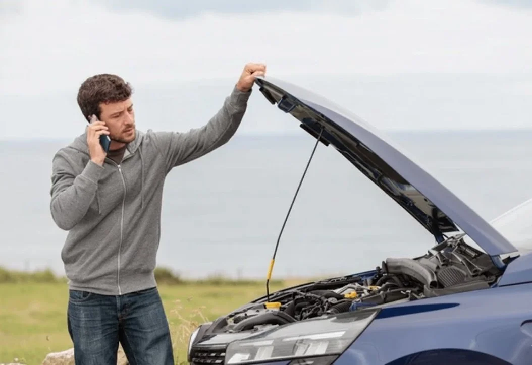 A man with a beard in a gray hoodie and jeans is standing outdoors, talking on a cell phone while inspecting a car with its hood open.