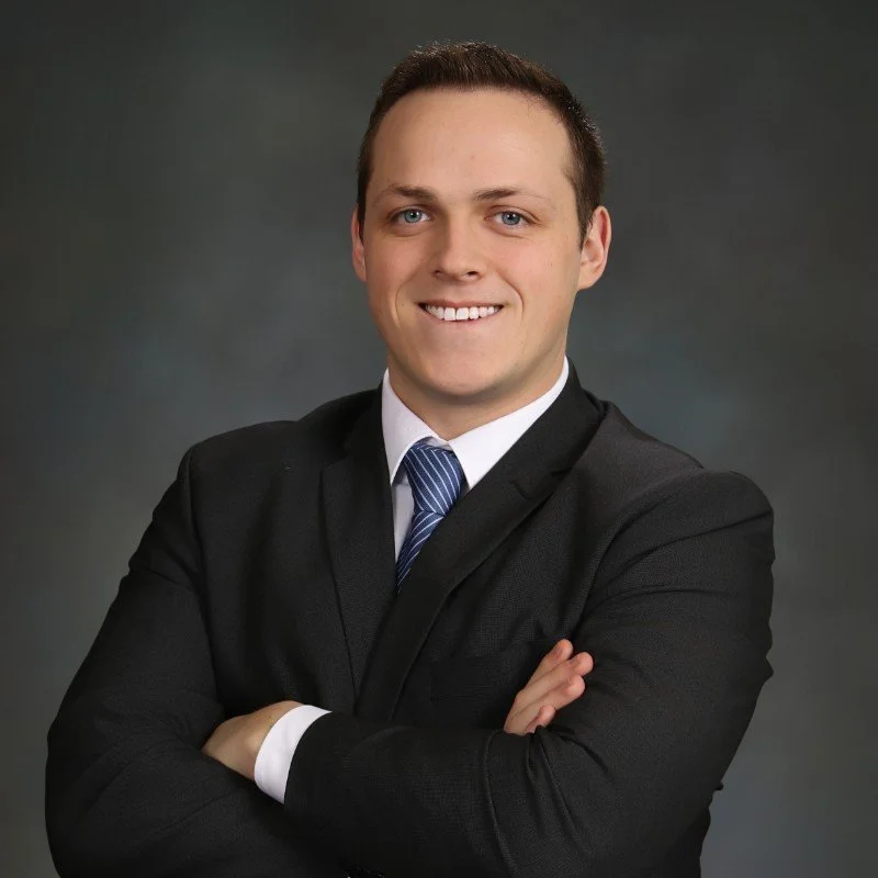 Professional young man in a black suit, white shirt, and blue striped tie, smiling with arms crossed against a dark gray background.