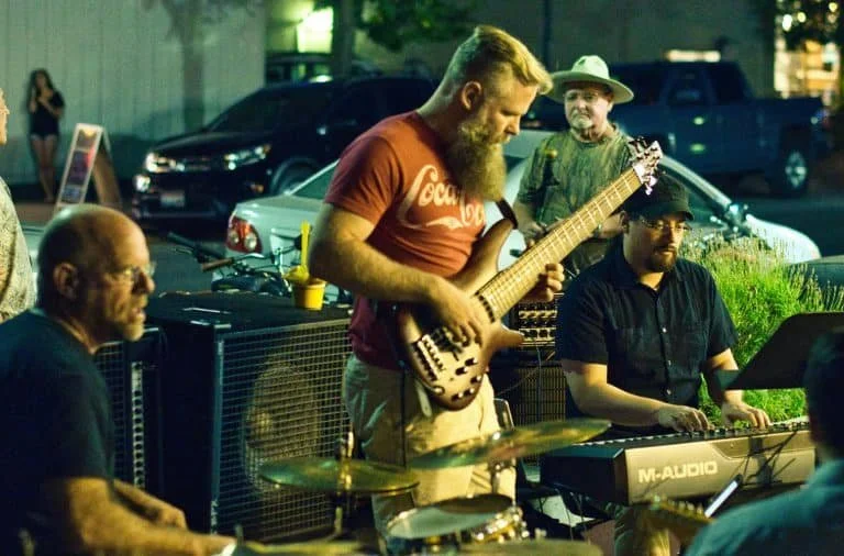 A group of musicians performing outdoors at night, including a guitarist, keyboard player, and drummer, with cars parked behind them.