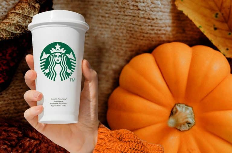 A hand holding a white Starbucks to-go cup with a green logo, placed beside a large orange pumpkin on a wooden surface surrounded by fall decorations.
