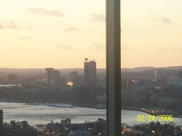 City skyline with buildings, river, and hills in the background, seen through a window divided by a vertical frame, with a timestamp reading 02/24/2006.