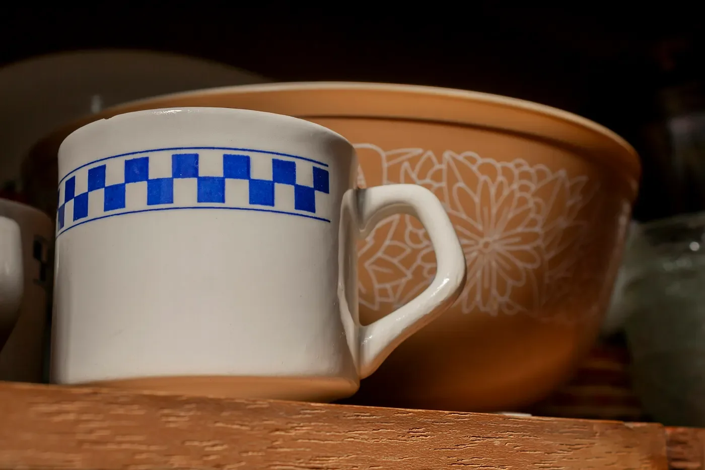 Close-up of a ceramic mug with a blue checkerboard pattern on a wooden shelf, with a larger orange bowl with white floral design behind it.