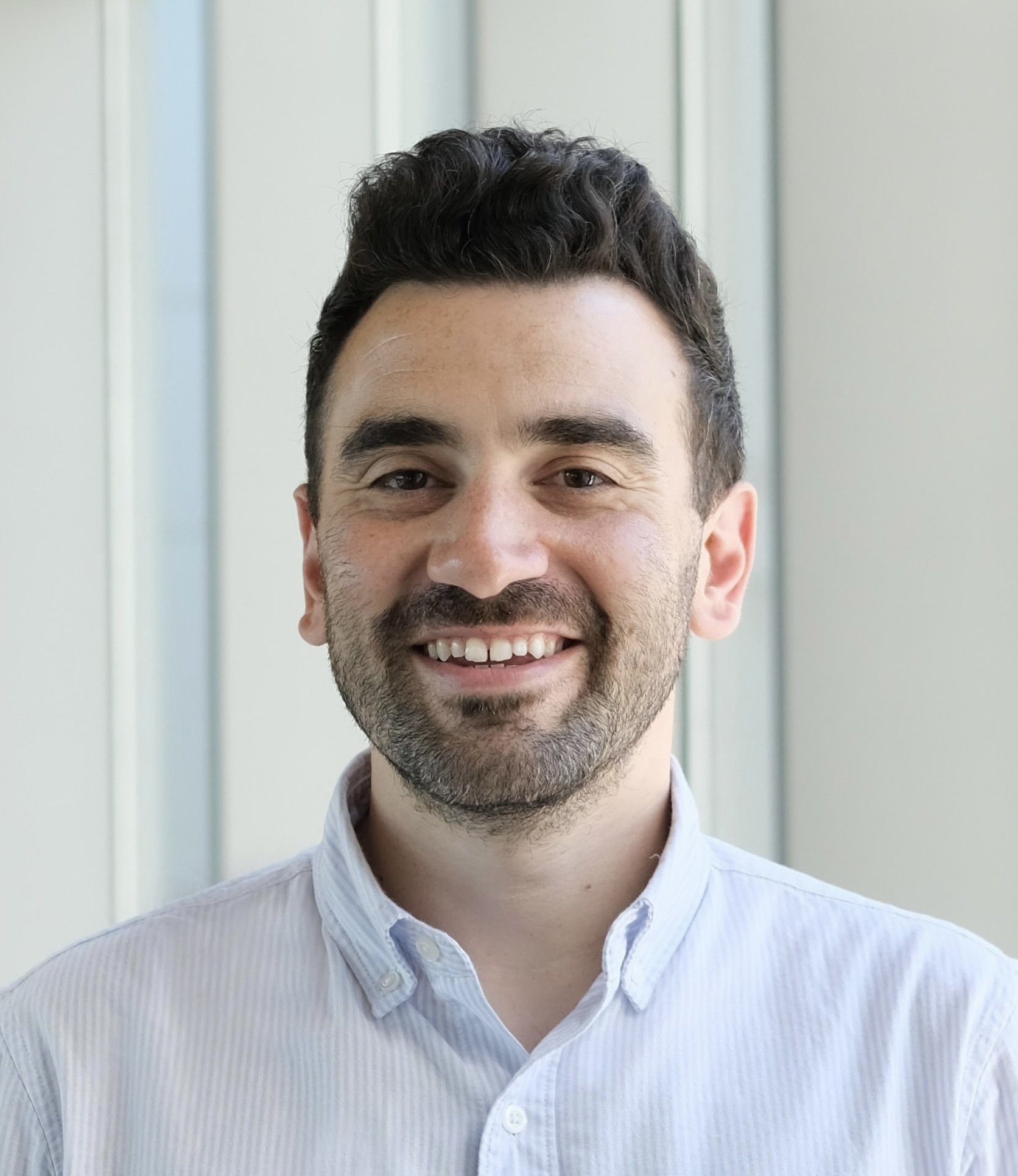Portrait of a smiling man with dark hair, a beard, wearing a white collared shirt, standing indoors near windows.