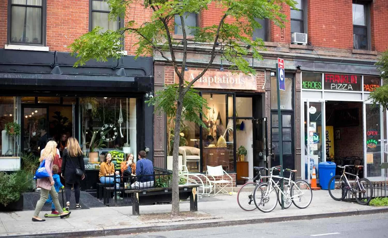 Street scene with a sidewalk, storefronts, bicycles, and people, including a girl riding a scooter and a group sitting outside a cafe.