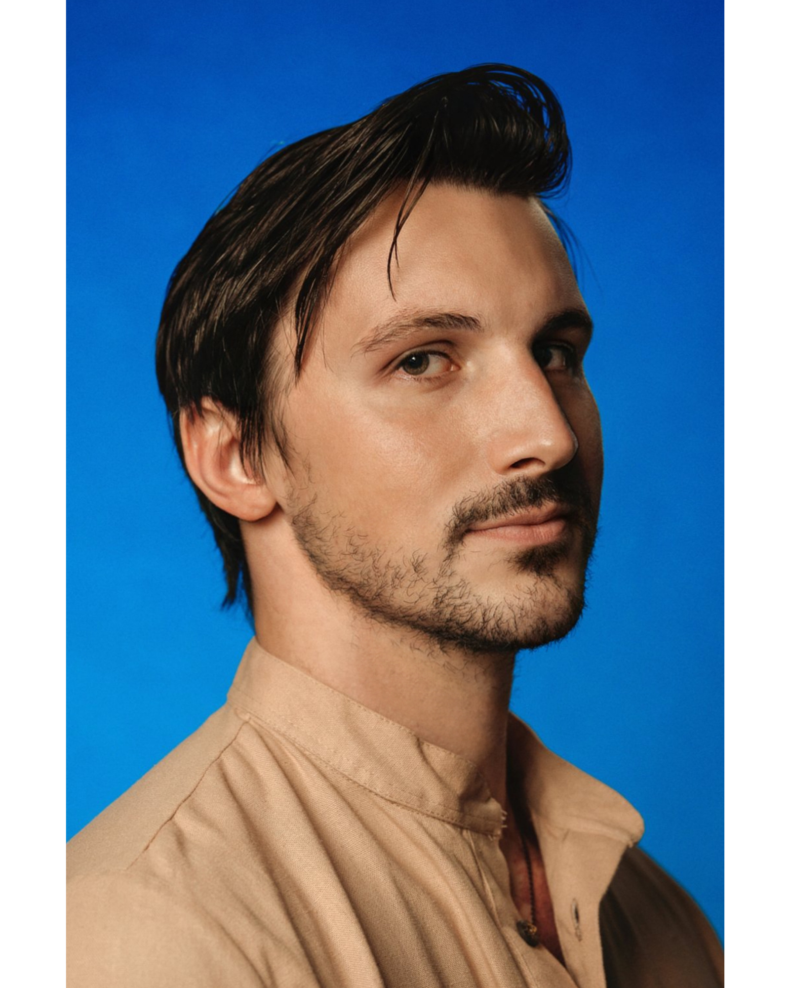Close-up portrait of a man with dark hair and facial hair, wearing a beige shirt, against a solid blue background.