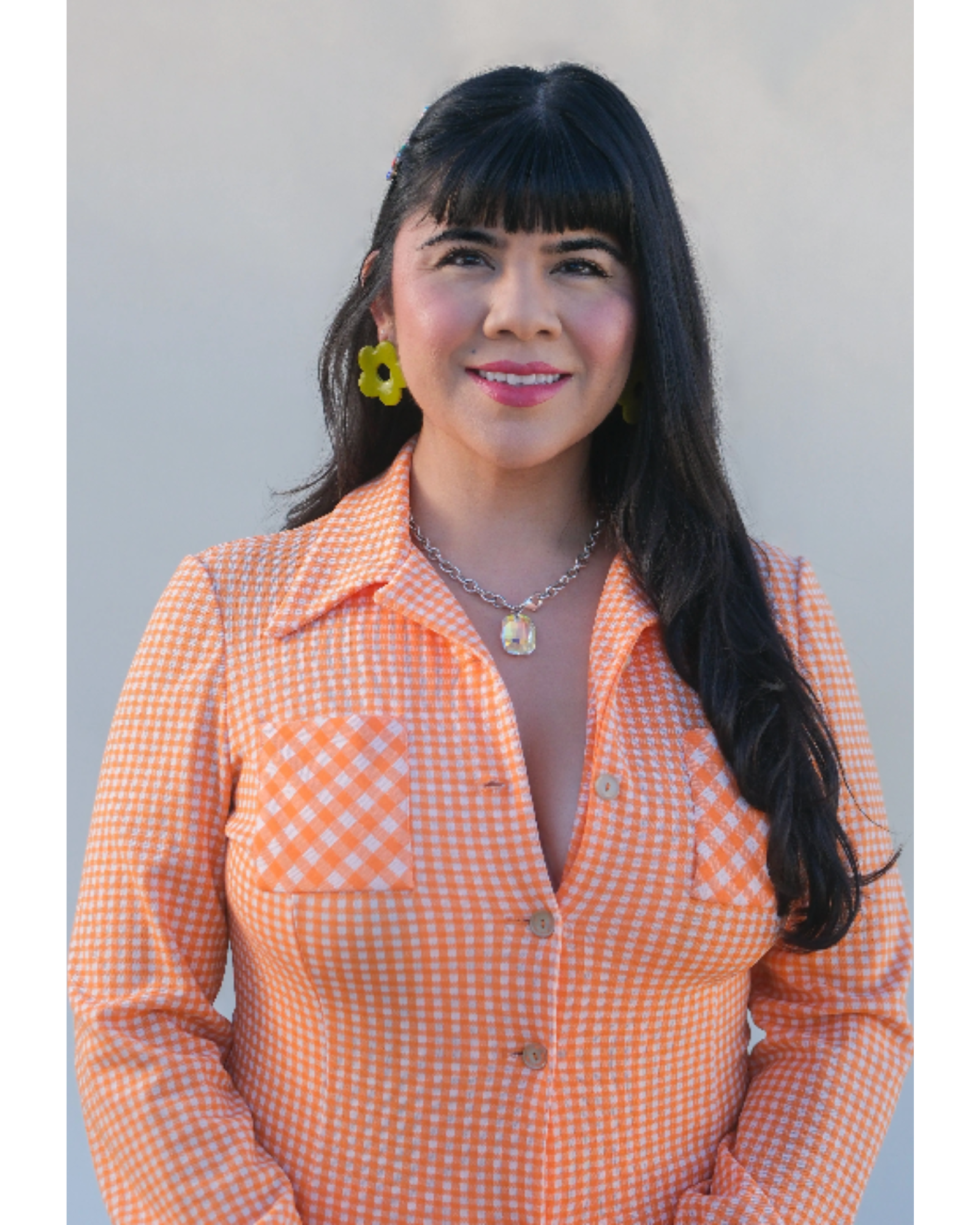 A woman with long black hair, wearing a orange checkered button-up shirt, yellow flower-shaped earrings, a silver necklace with a large gemstone, and standing against a plain background.